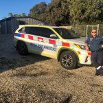 A paramedic stands in front of a MICA paramedic car. It's parked on gravel with a shed in the background. There is blue sky and the paramedic wears sunglasses.