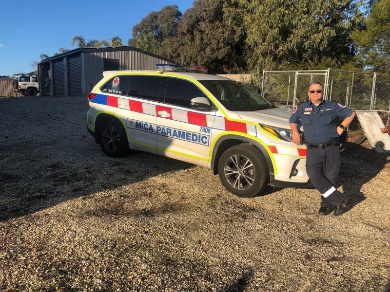 A paramedic stands in front of a MICA paramedic car. It's parked on gravel with a shed in the background. There is blue sky and the paramedic wears sunglasses.