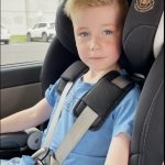 A young boy in a car seat in the back of a car, looking at camera