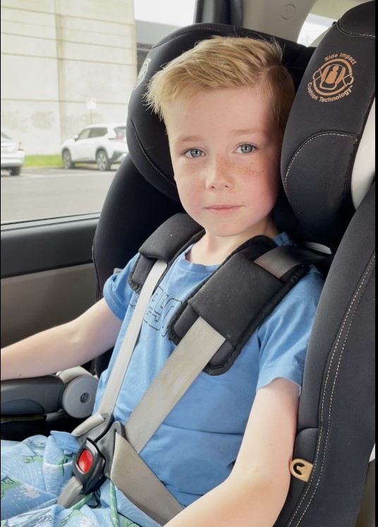 A young boy in a car seat in the back of a car, looking at camera