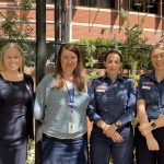 Four women smile for the camera. They're standing outside in front of a large brick building and a garden. Two wear paramedic uniform.