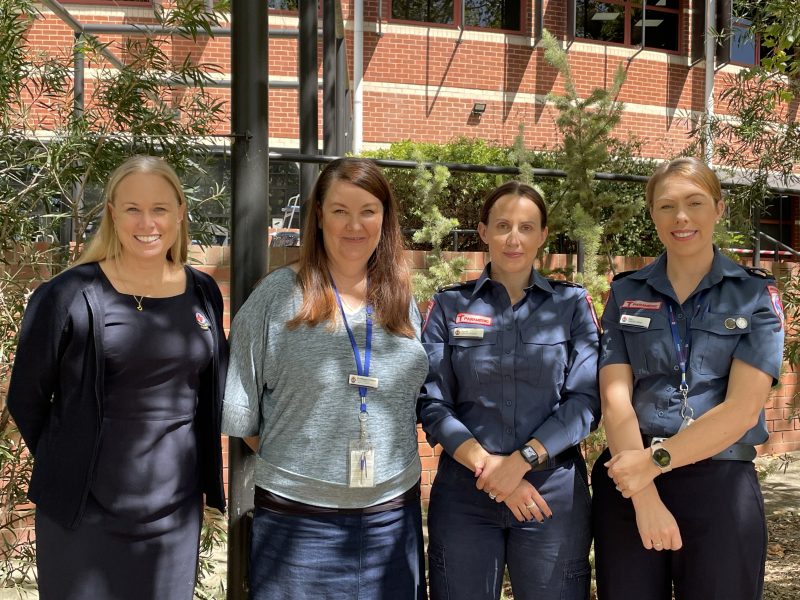 Four women smile for the camera. They're standing outside in front of a large brick building and a garden. Two wear paramedic uniform.