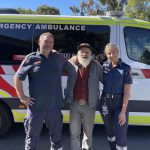 A man stands between two paramedics, who have their arms around him. They smile for the camera in front of an ambulance.