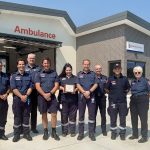 A group of people in Ambulance Victoria uniform smile for the camera in front of a modern ambulance branch. One woman holds a plaque.