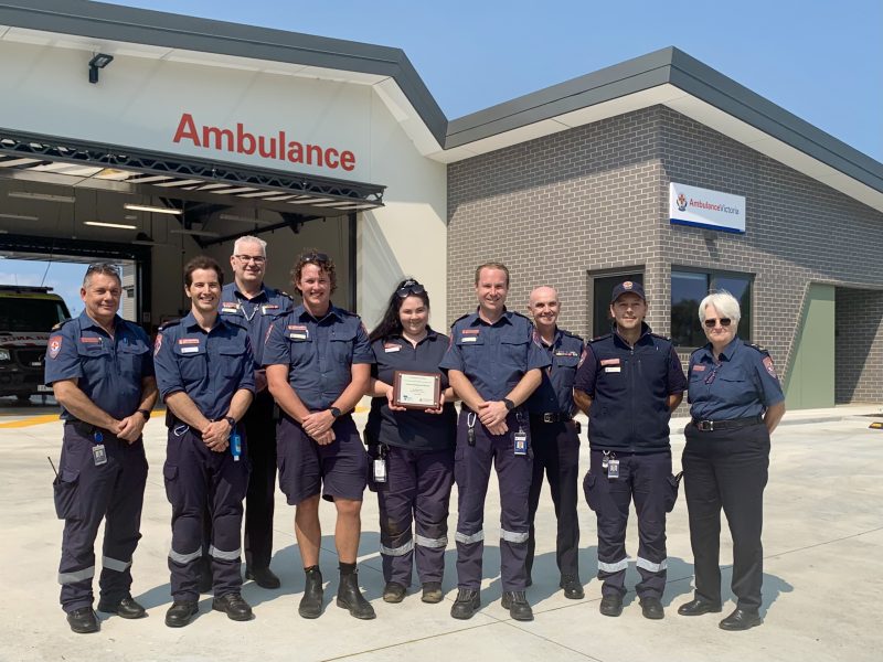 A group of people in Ambulance Victoria uniform smile for the camera in front of a modern ambulance branch. One woman holds a plaque.