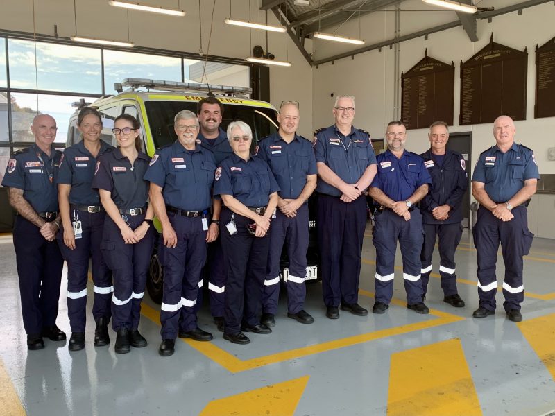 A group of people in Ambulance VIctoria uniform smile for the camera. They stand inside an ambulance branch garage, in front of a parked ambulance.