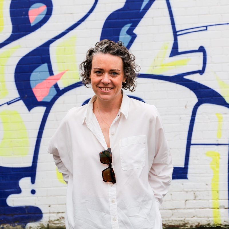 A woman wearing a white shirt smiles at the camera in front of a colourful, graffiti wall.