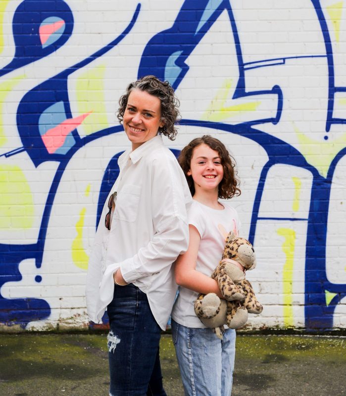 A woman and girl stand back-to-back in front of a colourful graffiti wall. The girl holds a toy animal.