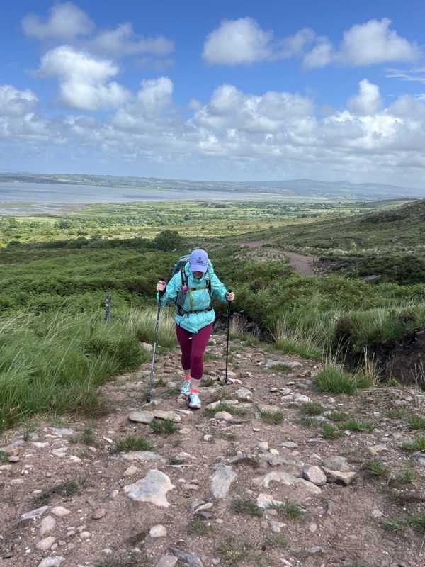 A woman hikes up a hill. It is a rocky path. Greenery spreads into the background and the coastline can be seen.