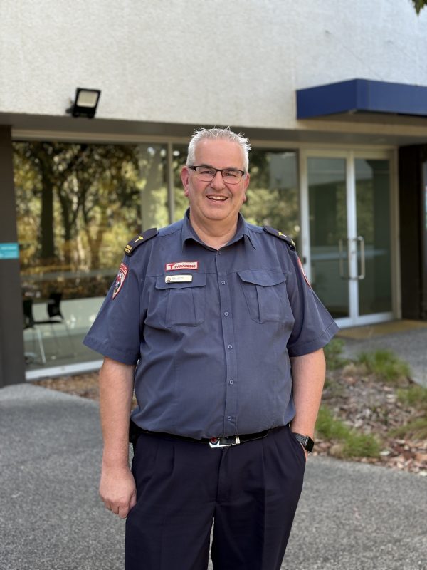 Paul in uniform, smiling and standing in front of an ambulance building