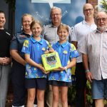 A group of adults and two young school students smile for the camera. The students hold an AED.