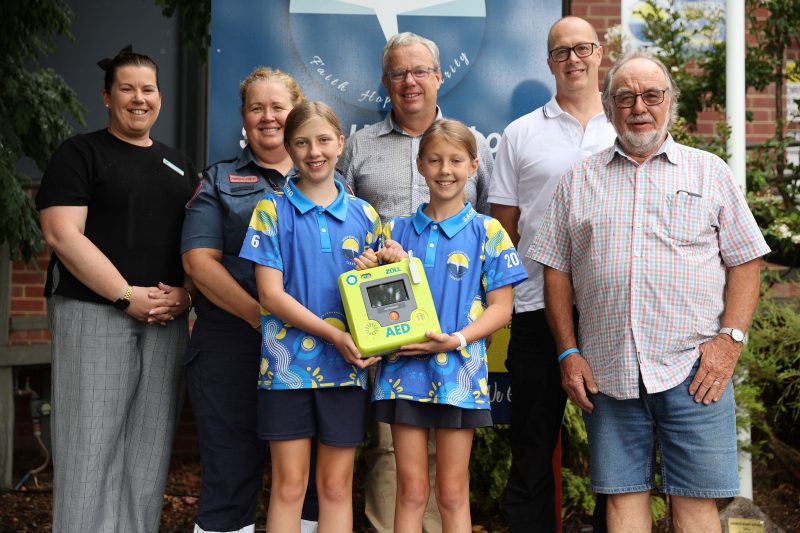 A group of adults and two young school students smile for the camera. The students hold an AED.