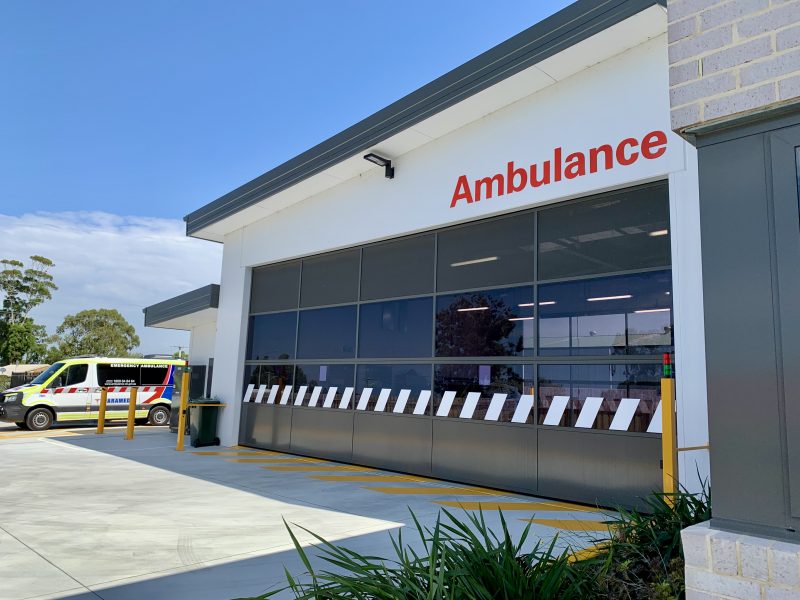 An ambulance branch garage door closed. An ambulance is parked next to it. The sky is blue.