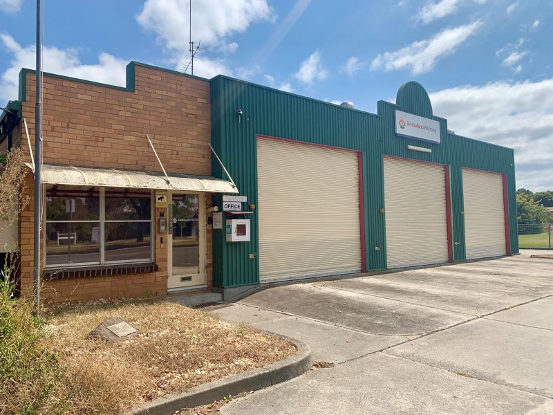 A small brick building is connected to a green garage with three roller doors. An Ambulance Victoria sign is above the roller doors.