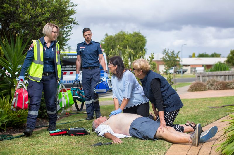 Two women help a man lying unconscious on the grass. Paramedics are arriving.